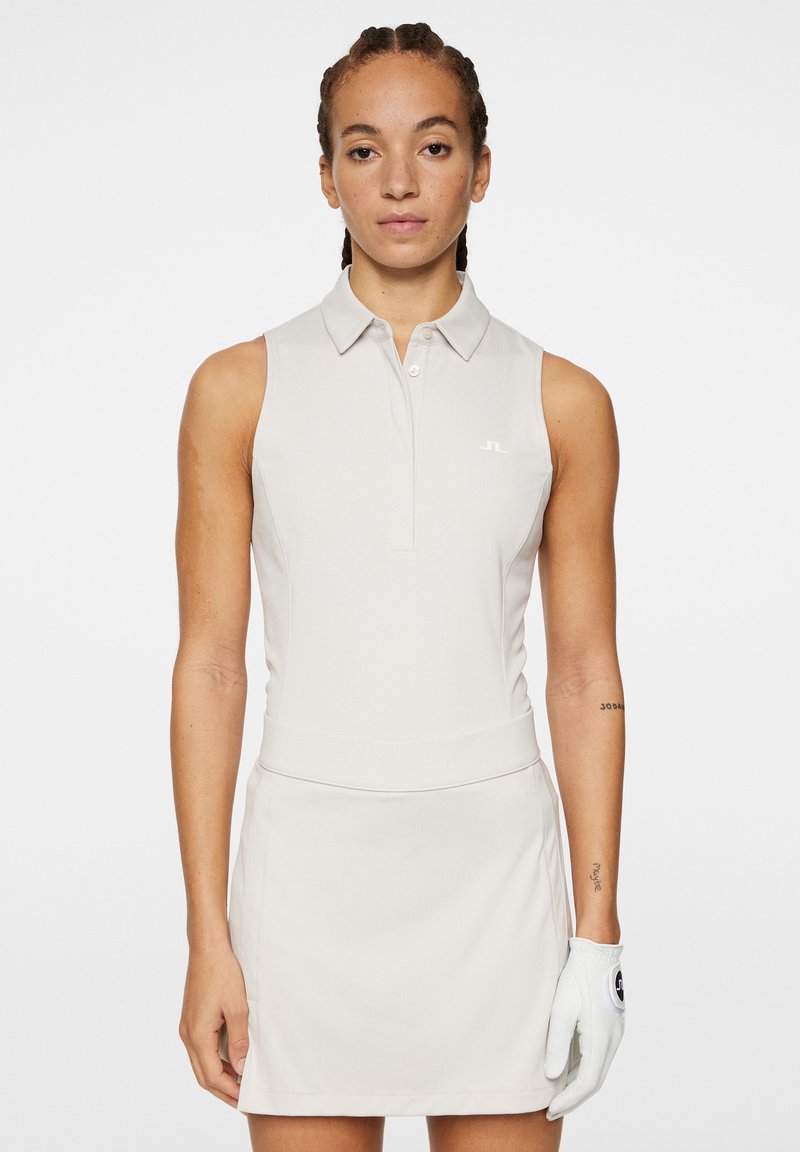 Ljusbeige ärmlös poloklänning med krage, figursydd midja och en slät yta. Bärs med en vit golfhandske.
