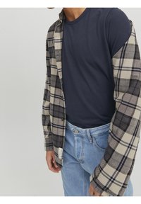 Man som bär en marinblå t-shirt under en öppen beige och svart rutiga skjorta tillsammans med ljusblå jeans mot en enfärgad bakgrund.