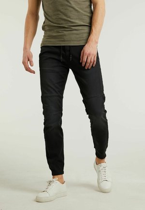 Homme portant un pantalon noir ajusté, une chemise à manches courtes vert olive et des baskets blanches, debout devant un fond clair uni.