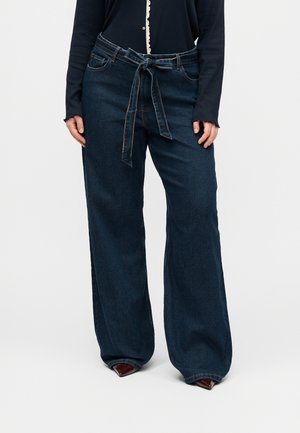 Person iført mørkeblå jeans med vide ben og et matchende denimbælte bundet om taljen samt spidse brune sko.