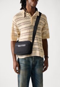Borsa a tracolla nera realizzata in materiale testurizzato, con un pannello frontale liscio e un logo. Indossata su una polo leggera a maglia con motivo.