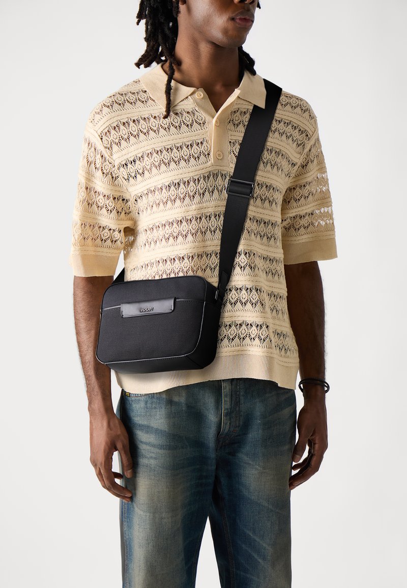 Borsa a tracolla nera realizzata in materiale testurizzato, con un pannello frontale liscio e un logo. Indossata su una polo leggera a maglia con motivo.