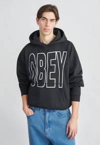 Fekete kapucnis pulóver nagy fehér "OBEY" felirattal, kenguru zsebbel, laza szabással és bordázott mandzsettákkal. Kék farmerrel párosítva.