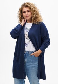 Cardigan long bleu marine, à manches longues, à l'avant ouvert, avec deux poches avant, texture côtelée, associé à un t-shirt graphique et un jean taille haute.