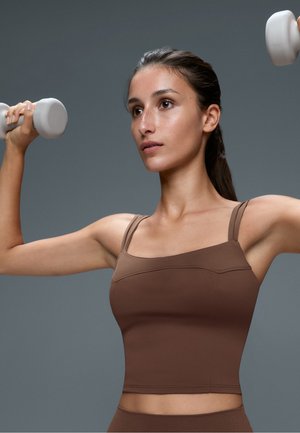 Giovane donna in abbigliamento sportivo marrone che solleva pesi grigio chiaro con entrambe le braccia davanti a uno sfondo grigio uniforme.