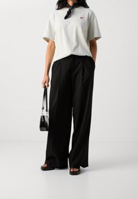 Jasnoszary bawełniany t-shirt z małym czerwonym logo, zestawiony z czarnymi spodniami o wysokim stanie i szerokimi nogawkami oraz czarnymi płaskimi sandałami, trzymający czarną torebkę.