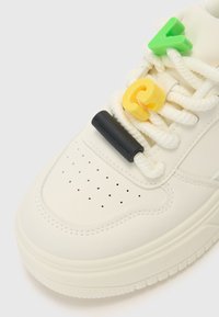 Weißer Sneaker mit einem glatten Obermaterial aus Leder. Besitzt bunte Kunststoffschnürsenkelclips in Grün, Gelb und Schwarz an dicken weißen Schnürsenkeln. Perforierte Zehenpartie.