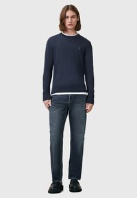 Dunkelblaues Sweater aus weichem Strick mit langen Ärmeln und Rundhalsausschnitt, über einem weißen Hemd getragen, kombiniert mit dunklen Jeans und schwarzen Schuhen.
