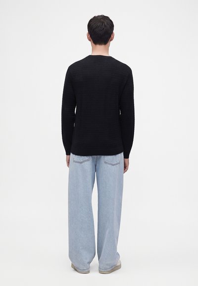 Homme portant une chemise noire à manches longues et un jean large bleu clair, debout de dos devant un fond blanc uni.