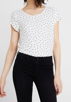 Kobieta w białej koszulce z krótkim rękawem z małym wzorem kotwic, włożona do czarnych spodni z wysokim stanem, z rękami swobodnie opuszczonymi wzdłuż ciała.