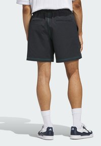 Schwarze Sportshorts mit elastischem Bund und türkisfarbenen Nähten; ausgestattet mit zwei Gesäßtaschen. Getragen mit weißen Socken und blauem Schuhwerk.