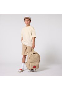 Μπεζ σακίδιο πλάτης με σχέδιο λογότυπου, που διαθέτει μπροστινή τσέπη και ταμπά διακοσμήσεις. Φοριέται με υφασμάτινο κρεμ πουκάμισο και μπεζ σορτς.