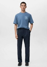 Niebieska bawełniana koszulka z nadrukiem na piersi, zestawiona z ciemnogranatowymi plisowanymi spodniami. Model stoi z rękami w kieszeniach.