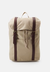 Sac à dos en toile beige avec des sangles en cuir marron, rabat repliable, fermeture à cordon et poche avant. Texture lisse, design minimaliste.