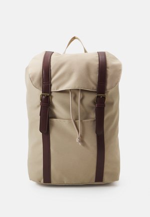 Sac à dos en toile beige avec des sangles en cuir marron, rabat repliable, fermeture à cordon et poche avant. Texture lisse, design minimaliste.