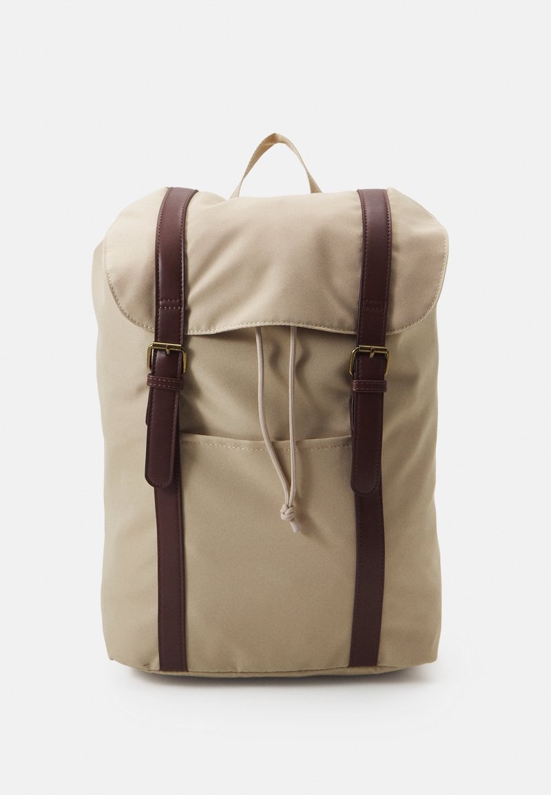 Beige canvas ryggsäck med bruna läderremmar, viklock, dragsko-stängning och framficka. Slät struktur, minimalistisk design.