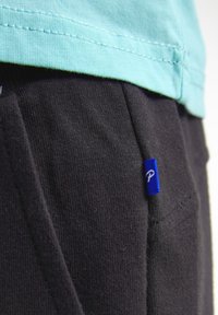 Lichtblauw katoenen t-shirt laag over zwarte trainingsbroek, met een klein blauw logo-label aan de zijkant en zichtbare stikdetails.