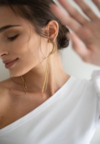 Gouden hoepel oorbellen met waterval kettingdetails, gedragen met een witte off-shoulder top. Zachte texturen en een gladde, glanzende afwerking.