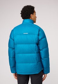 Veste en duvet teal avec des carrés rembourrés, col haut, et le logo "MAMMUT" en blanc au dos. Texture lisse, design léger.