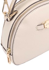 Coin de sac en cuir crème avec fermeture éclair en doré, fermoir et étiquette décorative en métal attachée au tirette de la fermeture.