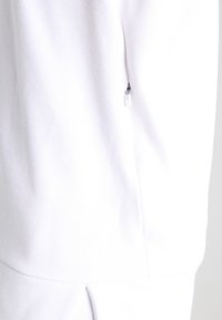 White garment showing a textured fabric with a side zipper detail. The zipper pull is clear plastic, blending with the fabric.