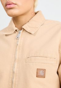 Giacca beige in denim, con zip frontale, collo a risvolto, tasca sul petto e una toppa quadrata in pelle con logo. Texture liscia.