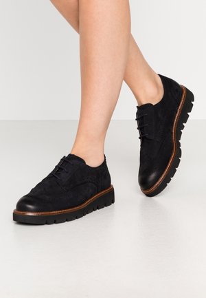 Sorte ruskindssko med en glat lædertå, detaljerede broguedekorationer, en struktureret sål og brune læderkanter langs bunden.