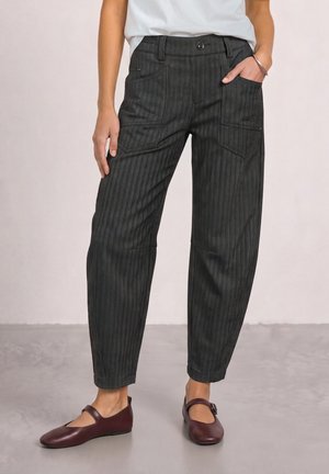 Femme portant un pantalon gris foncé à fines rayures avec des poches avant et des ballerines Mary Jane bordeaux, debout, une main dans la poche.