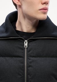 Black padded jacket with ribbed collar and silver zip closure. Smooth texture with horizontal paneling details. Minimalist design.