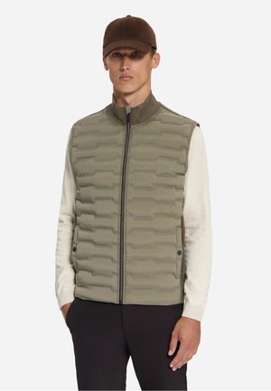 Man draagt een bruine pet, een beige overhemd met lange mouwen en een olijfgroene doorgestikte bodywarmer met rits en zakken, staand tegen een effen achtergrond.