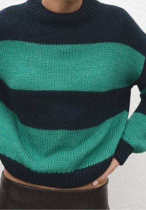 Personne portant un pull tricoté avec de larges rayures horizontales alternées bleu sarcelle et noir, avec un col rond, main reposant sur la hanche.
