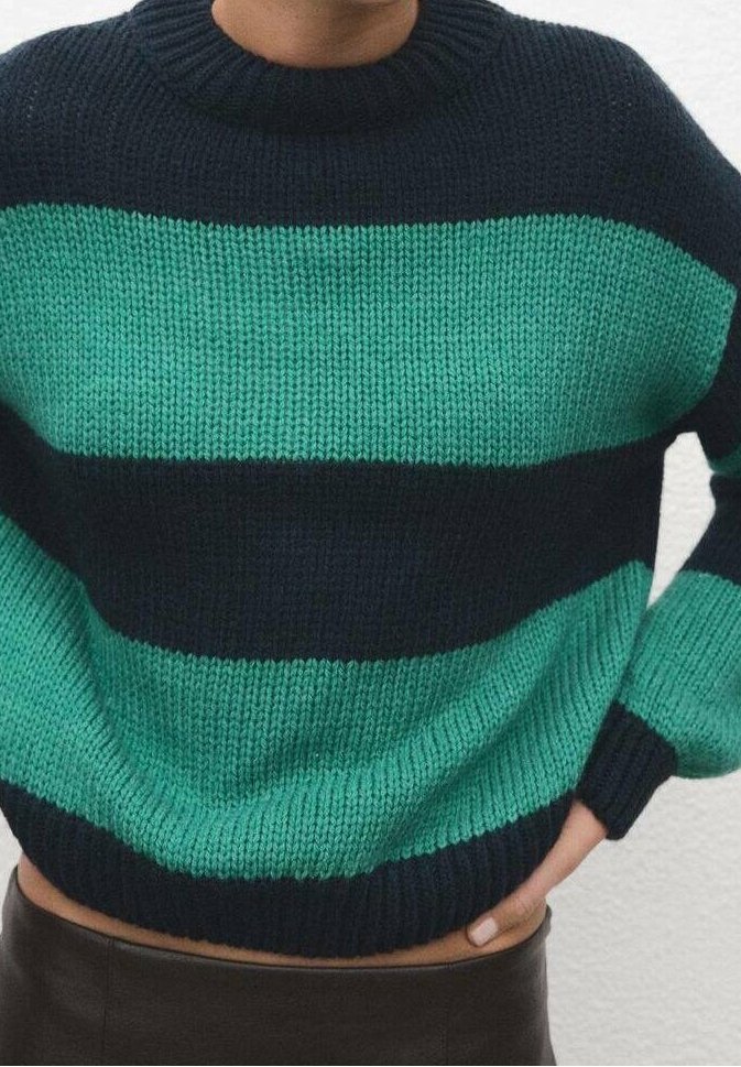 Personne portant un pull tricoté avec de larges rayures horizontales alternées bleu sarcelle et noir, avec un col rond, main reposant sur la hanche.