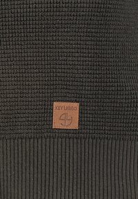 Der gestrickte dunkelgrüne Pullover hat einen strukturierten oberen Abschnitt und einen gerippten Saum. Ein braunes Leder-Patch zeigt das "KEY LARGO"-Logo.