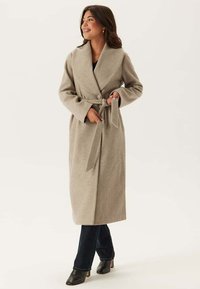 Helle beige lange Jacke mit Reverskragen, Taillengürtel und Knöpfen. Geschmeidige Textur, lange Ärmel und fällt unter die Knie, kombiniert mit dunklen Jeans.