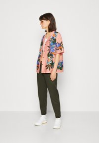 Chemise à manches courtes florale rose avec des motifs colorés, assortie à un pantalon vert olive et des baskets blanches. Modèle présenté en vue de profil.