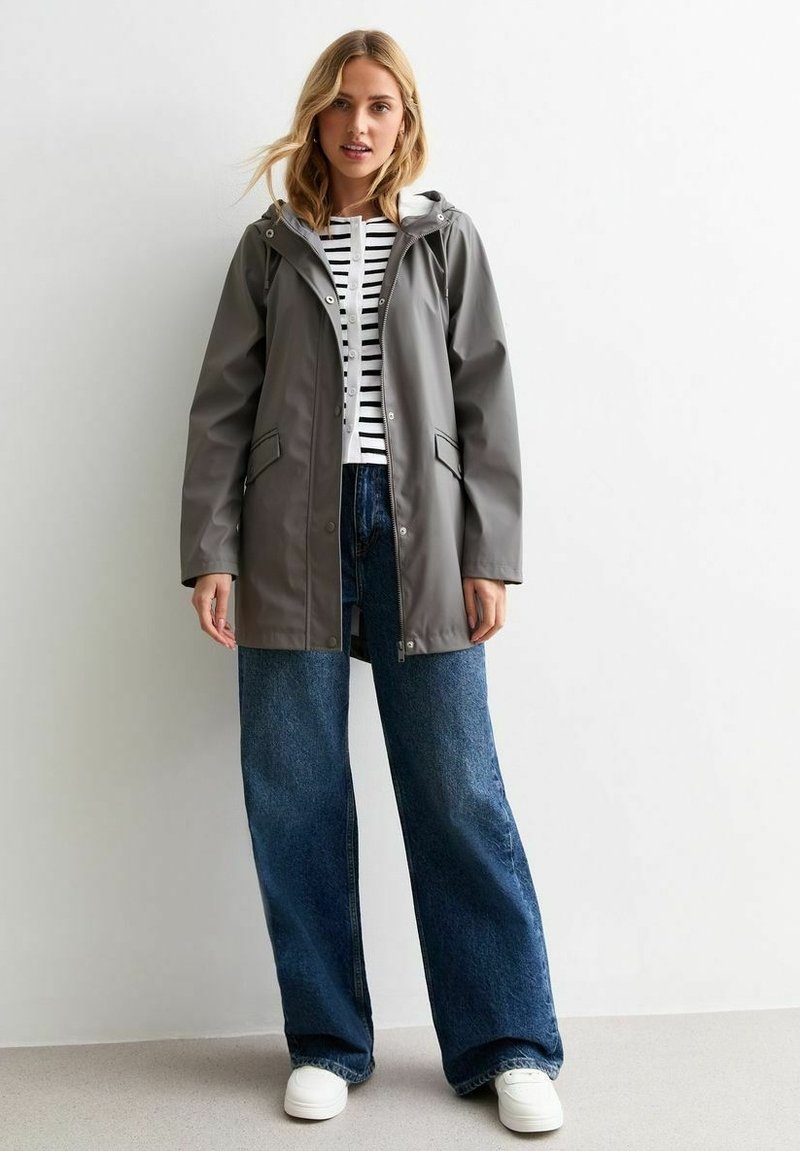 Impermeable de goma gris con capucha, cierres a presión y dos bolsillos, combinado con una camiseta de rayas blancas y negras y unos vaqueros de pierna ancha.