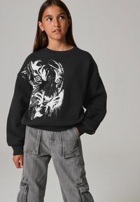 Sudadera negra con un gráfico de tigre blanco en el frente. Combinada con pantalones cargo grises, que muestran bolsillos y un ajuste relajado.