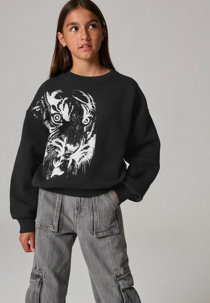 Sudadera negra con un gráfico de tigre blanco en el frente. Combinada con pantalones cargo grises, que muestran bolsillos y un ajuste relajado.