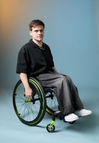 En person i svart skjorte og grå baggy bukser sitter i en svart rullestol med grønne detaljer, utstyrt med store hjul og et elegant design.