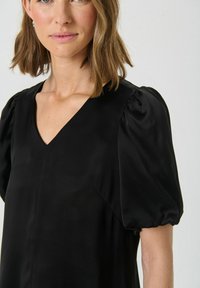 Rochie neagră cu decolteu în V, mâneci scurte puff și o textură netedă, lucioasă. Prezintă o siluetă simplă și detalii minime.