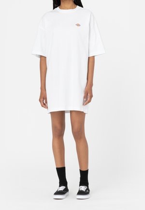 Kvinde iført en hvid, oversized T-shirt-kjole med en lille farverig logo på brystet, sorte sokker og sorte sneakers med hvide snørebånd.