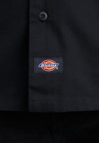 Maglietta nera realizzata in tessuto resistente, con chiusura a bottone e una colorata toppa con il logo "Dickies" vicino all'orlo.