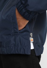 Casaco de nylon azul marinho com punhos e bainha elásticos, apresentando um bolso lateral com fecho éclair e um puxador prateado com um logótipo vermelho e amarelo.