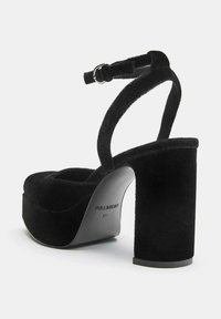 Mustat mokka-alustakengät paksuilla koroilla ja nilkkaremmillä. Ominaisuuksiin kuuluu pyöreä kärki ja sileä, matta pinta. Merkin logo näkyvissä pohjassa.