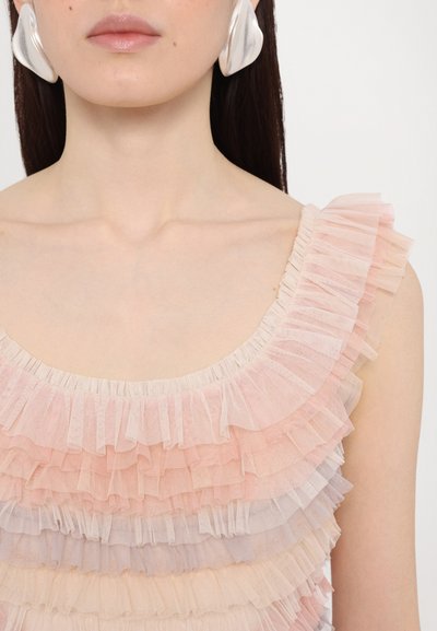 Roze gerimpelde top gemaakt van doorschijnende stof, met lagen in verschillende tinten roze en een gegolfde halslijn. Grote zilveren oorbellen accentueren de uitstraling.