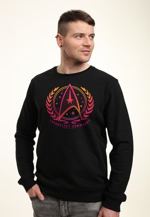 Homme portant un sweat-shirt noir avec un logo coloré de Starfleet Command, regardant sur le côté avec une main dans la poche sur un fond uni.
