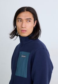Navy fleece jacket with high collar, zippered chest pocket in teal, and logo detail. Soft texture, relaxed fit, suitable for layering.