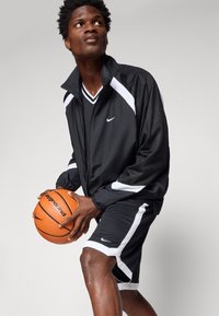 Junger Mann in schwarz-weißer Sportkleidung hält einen orangefarbenen Basketball und blickt aufmerksam zur Seite vor einem schlichten Hintergrund.