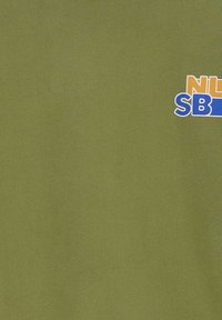 T-shirt en coton vert olive avec un design imprimé "NIL" en orange et "SB" en bleu. Texture lisse, coupe standard.