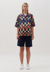 Mehrfarbiges Kurzarmhemd mit geometrischen Mustern in Pink, Blau, Grün und Beige, kombiniert mit marineblauen Shorts und weißen Sneakers.
